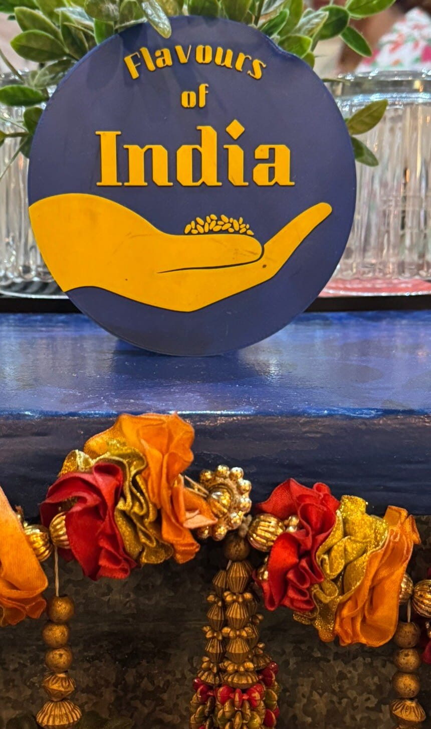 The Flavours of India badge standing on the blue counter shelf between jars and a flower garland